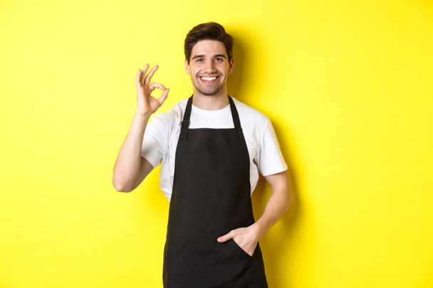 Jovem barista sorridente em avental preto mostrando sinal ok, recomendando café ou restaurante, de pé sobre fundo amarelo - Foto, Imagem