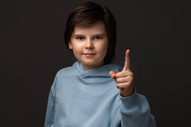Portrait of Serious boy 10-12 years old in casual clothes showing index finger, scolding or telling off someone. Little boy raises finger up, says: Attention please. Studio shot, gray background - Photo, Image
