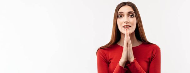 Portret van vrolijke brunette vrouw met lang haar in casual rode trui die handpalmen bij elkaar houdt en bidt. Ze mediteert, bidt voor vrede en liefde, heeft een rustige en vredige gelaatsuitdrukking - Foto, afbeelding