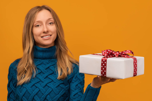 Imagen de la encantadora mujer joven en suéter de punto cálido sonriendo y sosteniendo regalo con cinta roja. Fotografía del estudio, fondo amarillo. Año Nuevo, Día de la Mujer, Cumpleaños, Concepto de vacaciones - Foto, imagen