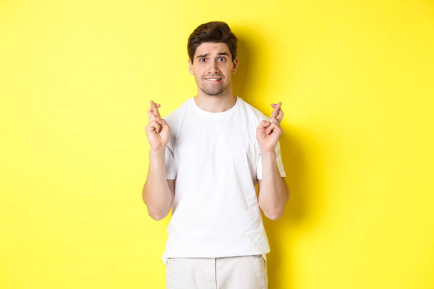 Nerwowy facet trzymający kciuki na szczęście, z nadzieją na coś, stojący nad żółtym tłem - Zdjęcie, obraz