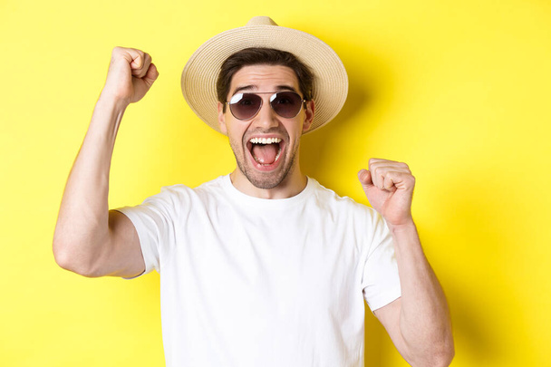 Concept of tourism and holidays. Happy male tourist celebrating his vacation, raising hands up and shouting for joy, wearing sunglasses with straw hat - Photo, Image