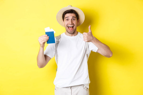 Tourism and vacation. Satisfied male tourist showing passport with tickets and thumb up, recommending travel company, standing over yellow background - Photo, Image