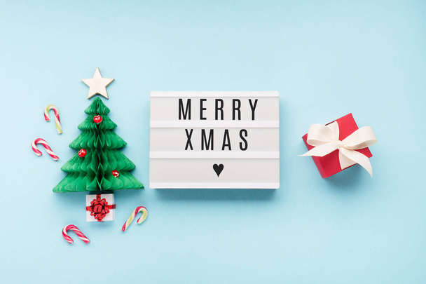 Merry Xmas text on white Lightbox with Christmas paper green tree, mushrooms and red gift box on blue background. Flat lay, top view, copy space. Festive holiday card concept. - Photo, image