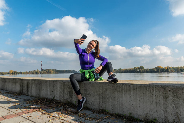 Young handsome sporty active young woman in sports wear sitting outdoor and taking a selfie with hear smartphone after recreational exercise and taking a break from workout training - Zdjęcie, obraz