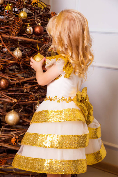 Egy elegáns fehér és arany ruhás kislány áll háttal a kamerának, és díszíti a karácsonyfát. Függőleges fénykép - Fotó, kép