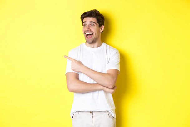 Homme souriant excité pointant et regardant à gauche, regardant promo offre, debout sur fond jaune - Photo, image