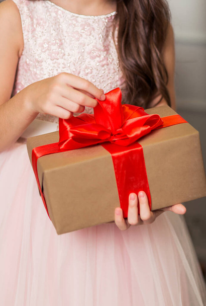 Uma menina em um vestido chique fica com um presente de Ano Novo em suas mãos contra o fundo de uma árvore de Ano Novo. Foto vertical - Foto, Imagem