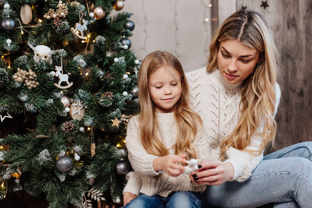 Young mother and her cute daughter sitting near Christmas tree indoors. - Φωτογραφία, εικόνα