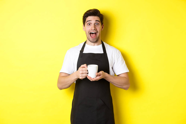 Young male barista holding coffee cup and looking surprised, standing in black apron against yellow background - Photo, Image
