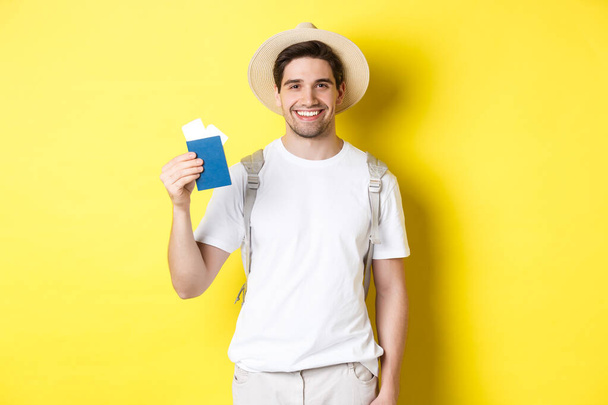 Tourism and vacation. Young smiling tourist showing passport with tickets, going on a trip, standing against yellow background - Fotoğraf, Görsel