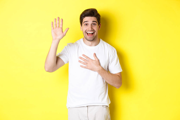 Happy guy making promise, holding hand on heart, swearing to tell truth, standing over yellow background in white t-shirt - Foto, Bild