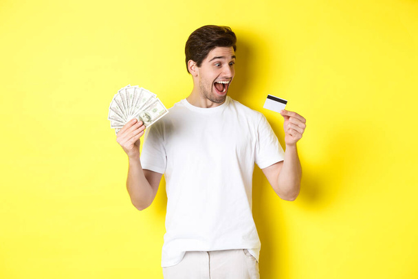 Kredi kartına bakan neşeli adam, elinde para, banka kredisi ve kredi konsepti, sarı arka planda duruyor. - Fotoğraf, Görsel