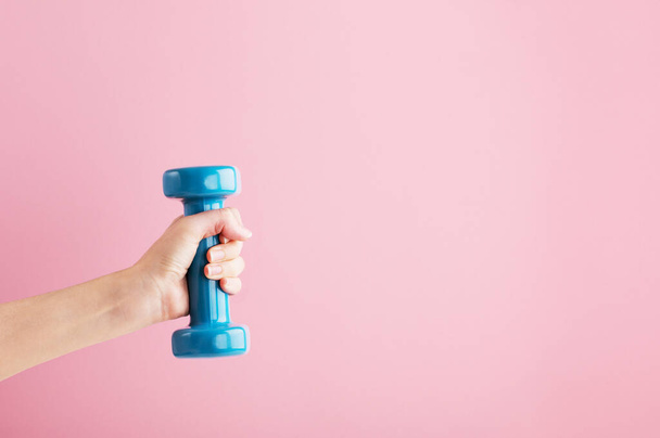 Persons hand lifting blue dumbbell. Fitness equipment for home and gym workout. Train indoors. Sport and healthy lifestyle concept. Copy space in right side. Isolated on pink background. - Photo, Image