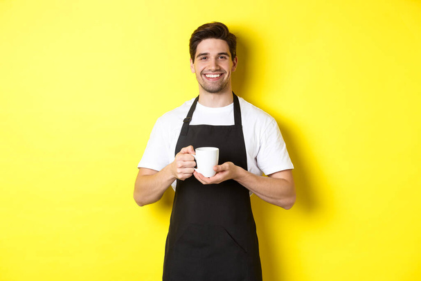 Bello barista che serve caffè, portare tazza, in piedi in grembiule nero con sorriso amichevole - Foto, immagini