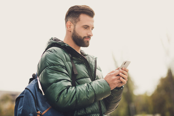 Profile side photo of good mood positive young man wear green windbreaker schoolbag texting modern device outside city street - 写真・画像