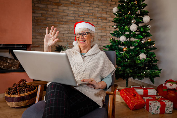 Joyeux rire femme âgée à la maison, avec ordinateur ayant un appel vidéo et agitant les mains à la maison. Noël, vacances, communication en ligne, distance sociale, isolement personnel, concept de quarantaine. - Photo, image