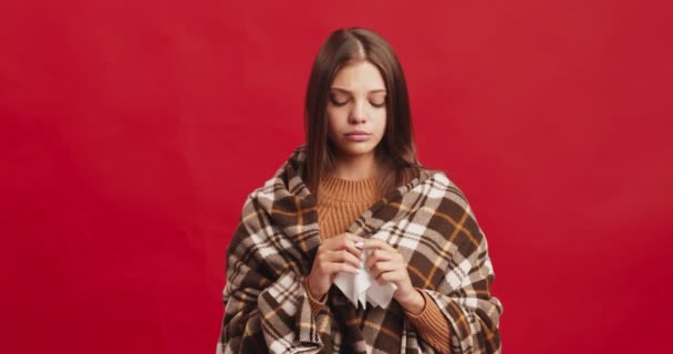 Νεαρή κοπέλα τυλιγμένη σε καρό φτάρνισμα, φυσώντας τη μύτη της σε ιστό, αρρώστησε πριν τις γιορτές, κόκκινο φόντο στούντιο - Πλάνα, βίντεο