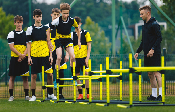 Chłopiec skaczący przez płotki na treningu piłki nożnej. Drużyna piłki nożnej na treningu z młodym trenerem. Młodzież w klasie wychowania fizycznego - Zdjęcie, obraz