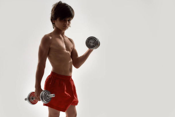 Little sportive boy child with muscular body looking at camera, showing his muscles, lifting weights while standing isolated over white background - Photo, Image