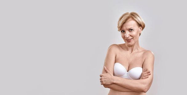 Attractive middle aged woman in underwear looking at camera while posing, standing against grey background - Foto, afbeelding