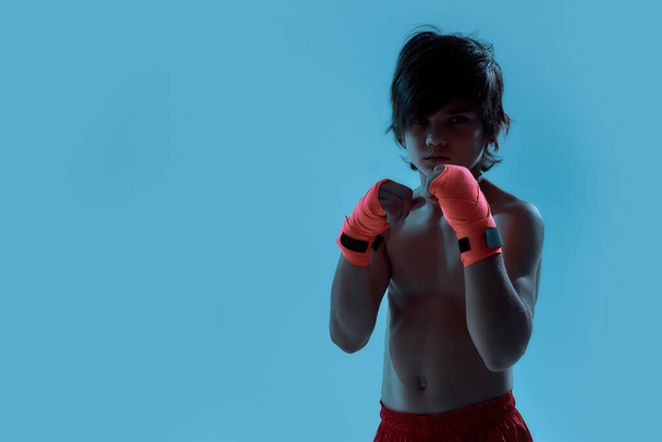 Silhouetted shot of little boy boxer in shorts with boxing bandage on hands, looking at camera ready to fight while posing, standing isolated over blue background - Fotografie, Obrázek