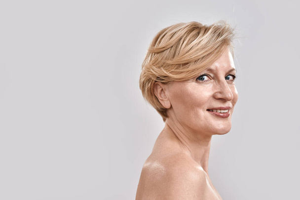 Portrait of beautiful naked middle aged woman smiling at camera while posing isolated against grey background. Beauty, skincare concept - Foto, Imagem