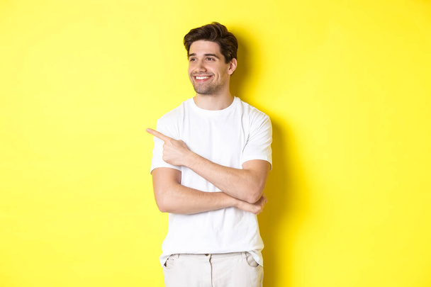 bel homme souriant dans des vêtements blancs, regardant et pointant du doigt gauche à la bannière, debout sur fond jaune - Photo, image