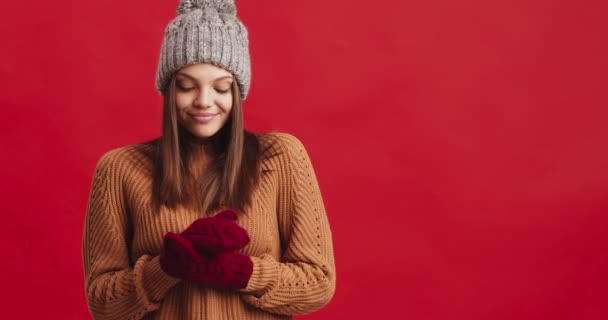 Iloisen naisen muotokuva talvihatussa ja lapasissa jäätyi, lämpeni ja hymyili, punainen tausta - Materiaali, video
