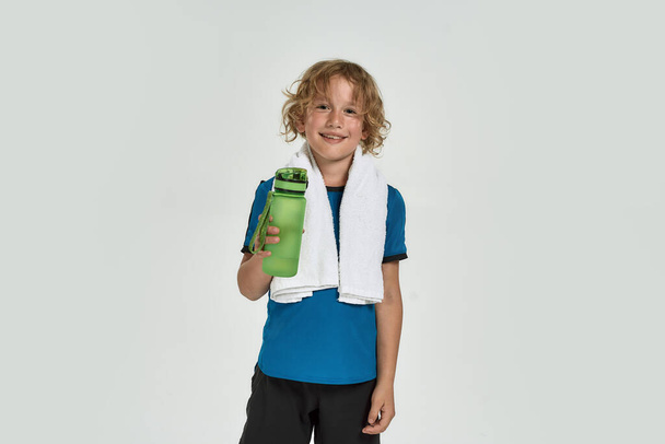 Portrait of little sportive boy child in sportswear smiling at camera, holding water bottle while standing with towel around his neck isolated over white background - Photo, Image