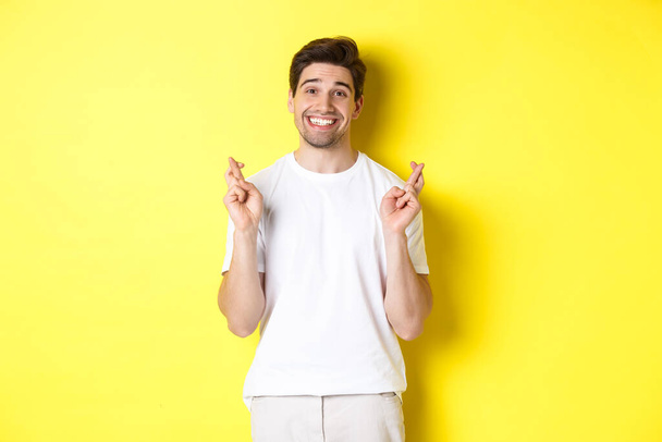 Hopeful smiling man holding fingers crossed, making wish or praying, standing over yellow background - Фото, изображение