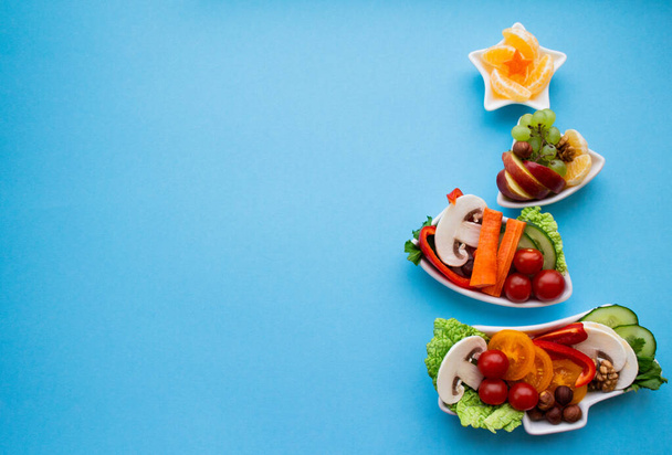Bord in de vorm van een kerstboom met groenten, fruit, champignons en bessen. Blauwe achtergrond. Het concept van vegetarische lekkernijen voor de vakantie. Kopieerruimte. - Foto, afbeelding