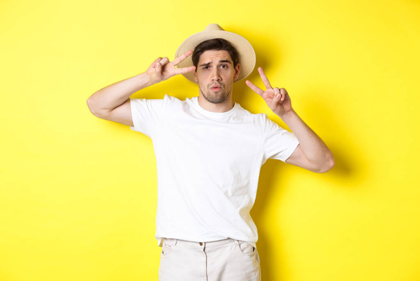 Pojęcie turystyki i wakacji. Fajny facet robiący zdjęcia na wakacjach, pozujący ze znakami pokoju i noszący słomiany kapelusz, stojący na żółtym tle - Zdjęcie, obraz