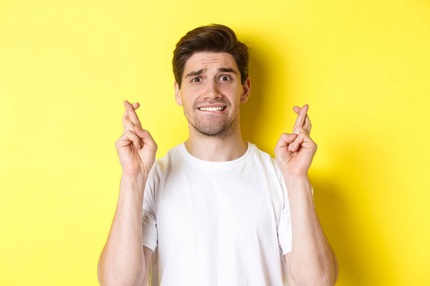 Κοντινό πλάνο του νευρικού άνδρα που κάνει ευχή, κρατώντας τα δάχτυλα σταυρωμένα και δαγκώνοντας τα χείλη ανήσυχοι, στέκεται πάνω από κίτρινο φόντο - Φωτογραφία, εικόνα