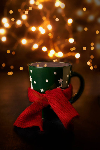 christmas scene. decorated mug of cocoa and red scarf around the mug. And festive lights on the background - Photo, Image