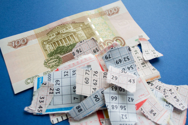 Москва, Россия, июль 2020: порванные лотерейные билеты с номерами с банкнотой в сто рублей на синем фоне. Русский лото. - Фото, изображение