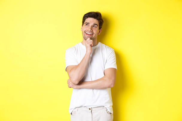 Portret uśmiechniętego mężczyzny wyglądającego troskliwie w lewym górnym rogu, wybierającego coś, mającego pomysł, stojącego na żółtym tle - Zdjęcie, obraz