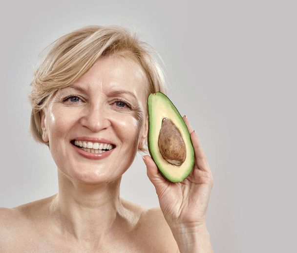 Portait of beautiful middle aged woman smiling at camera while holding half of a ripe delicious avocado near her face, posing isolated over grey background - Photo, Image