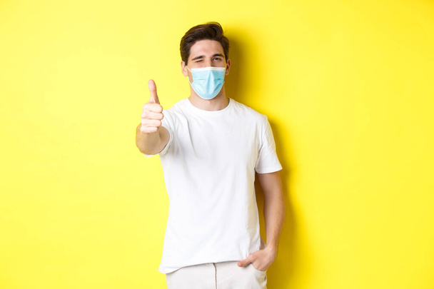 Koncept koronaviru, pandemie a sociální distancování. Sebevědomý mladý muž v lékařské masce ukazující palce nahoru a blikající, žluté pozadí - Fotografie, Obrázek