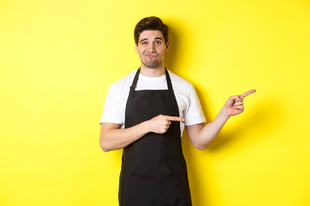 Kétséges barista fekete kötényben ujjal mutogatva jobbra, szkeptikusnak és szórakoztatatlannak tűnik, sárga háttér felett áll - Fotó, kép