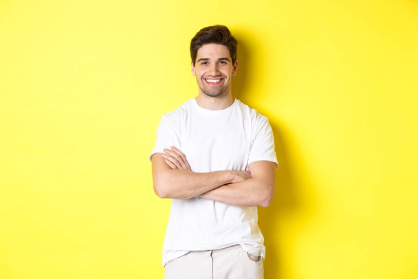 Wizerunek pewnego siebie białego mężczyzny uśmiechającego się zadowolonego, trzymającego się za ręce skrzyżowane na klatce piersiowej i wyglądającego na zadowolonego, stojącego nad żółtym tłem - Zdjęcie, obraz