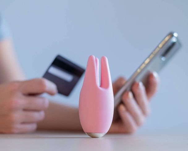 Faceless woman holding a credit card smartphone. Clitoral vibrator on the table. Online sex shop concept. - Fotó, kép