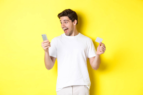 Surprised guy holding smartphone and credit card, online shopping on black friday, standing over yellow background - Фото, изображение