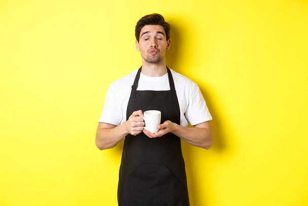 Hombre en delantal negro trayendo taza de café y esperando un beso, de pie sobre fondo amarillo - Foto, imagen