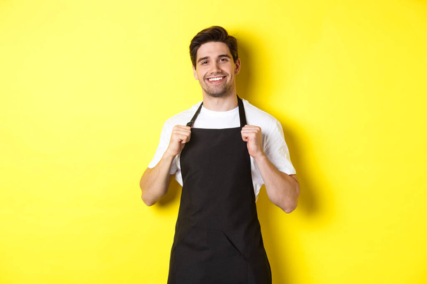 Sebevědomý barista v černé zástěře, stojící na žlutém pozadí. Číšník se usmívá a vypadá šťastně - Fotografie, Obrázek