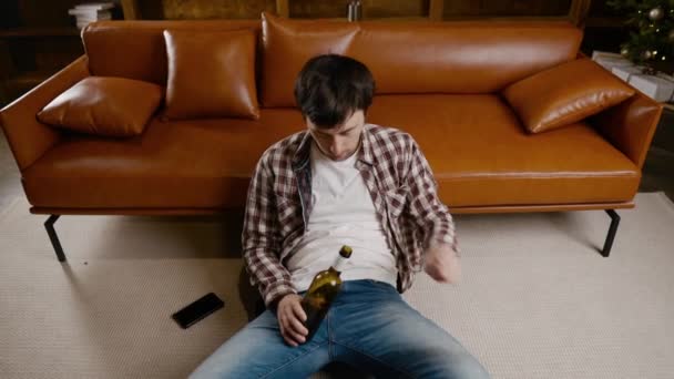 Joven deprimido bebiendo alcohol en casa. Hombre desempleado, borracho y sigue bebiendo vino botella recta en el sofá cerca del árbol de Navidad. Adicción, soledad, nerviosismo año nuevo vacaciones - Metraje, vídeo