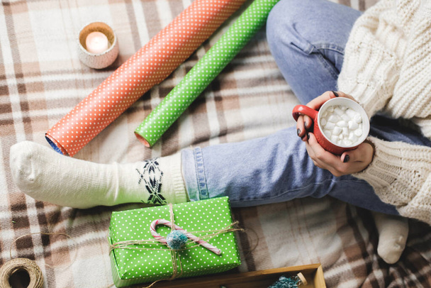 Young woman sits on plaid in cozy knitted woolen white sweater and holds cup of cocoa with marshmallows. Hygge New Year, preparation for holidays. Candles, Christmas balls, gifts, wrapping paper. - Foto, immagini