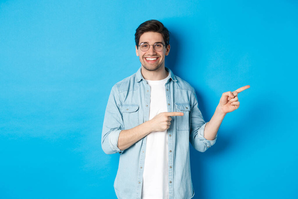 Bonito modelo masculino caucasiano em óculos, apontando os dedos para o seu logotipo, mostrando espaço de cópia, em pé sobre fundo azul - Foto, Imagem
