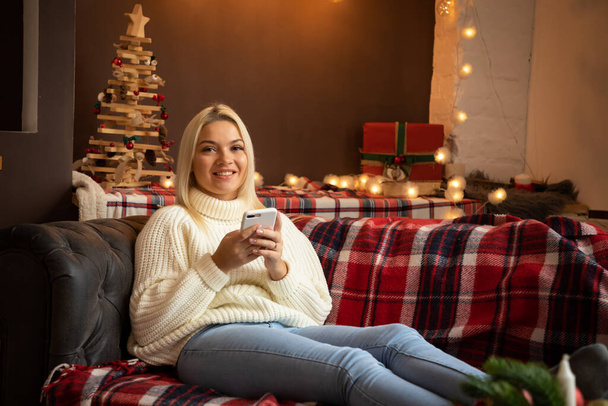 クリスマス休暇でソファに座っている間に家で幸せな女性はテキストメッセージを読んで、幸せな新年 - 写真・画像