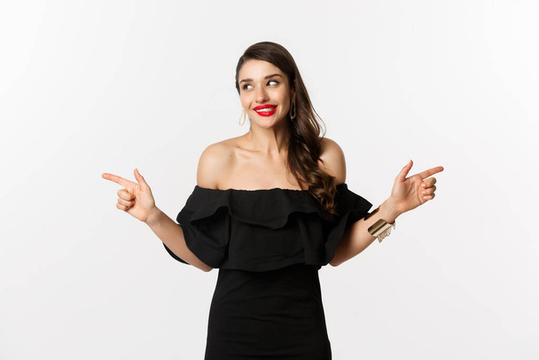 Fashion and beauty. Attractive woman in jewelry, makeup and black dress, smiling and pointing fingers sideways copy space offer, white background - Fotografie, Obrázek